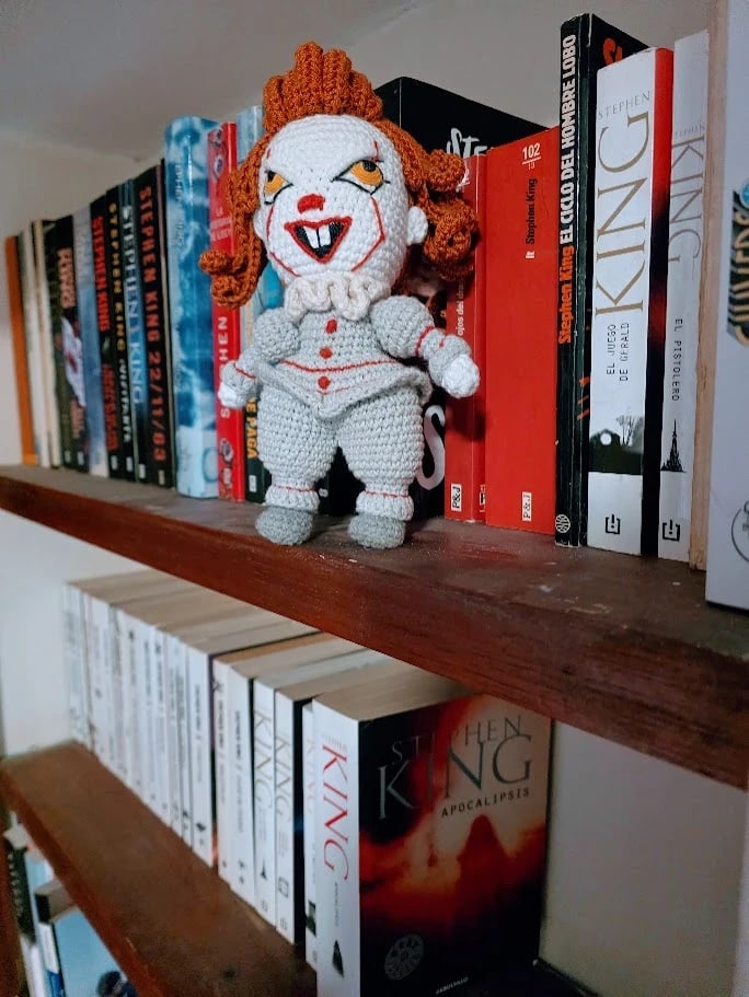 Un Pennywise tejido a crochet, de pie sobre un estante con libros de Stephen King; a la derecha asoma El pistolero y en el estante de abajo, Apocalipsis.